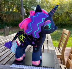 Size: 2048x1962 | Tagged: safe, artist:orky, derpibooru import, oc, oc only, oc:twinkle pop, pony, unicorn, black sclera, book, chair, commission, female, horn, irl, leonine tail, lidded eyes, mare, outdoors, photo, plushie, pony plushie, smiling, solo, standing, table, tail, unicorn oc
